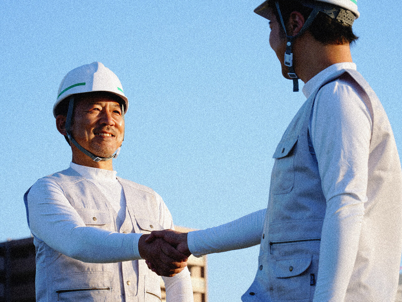 ヘルメットを着用した建設作業員同士が青空の下で握手する様子
